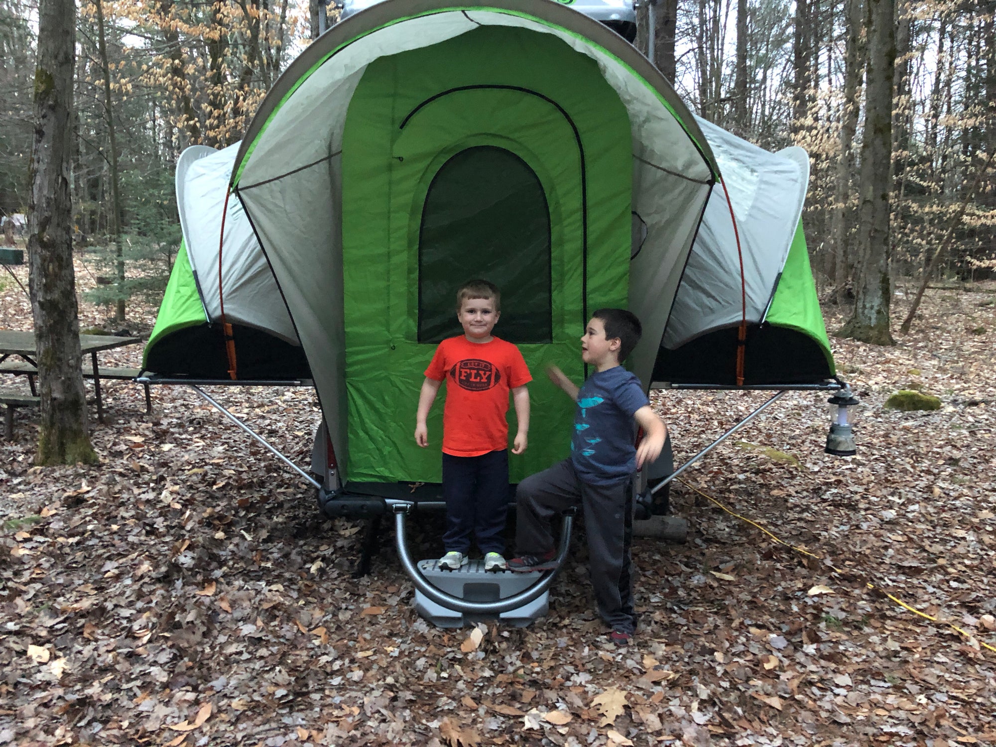 2 Boys stand in front of green unconventional tent structure in the woods.
