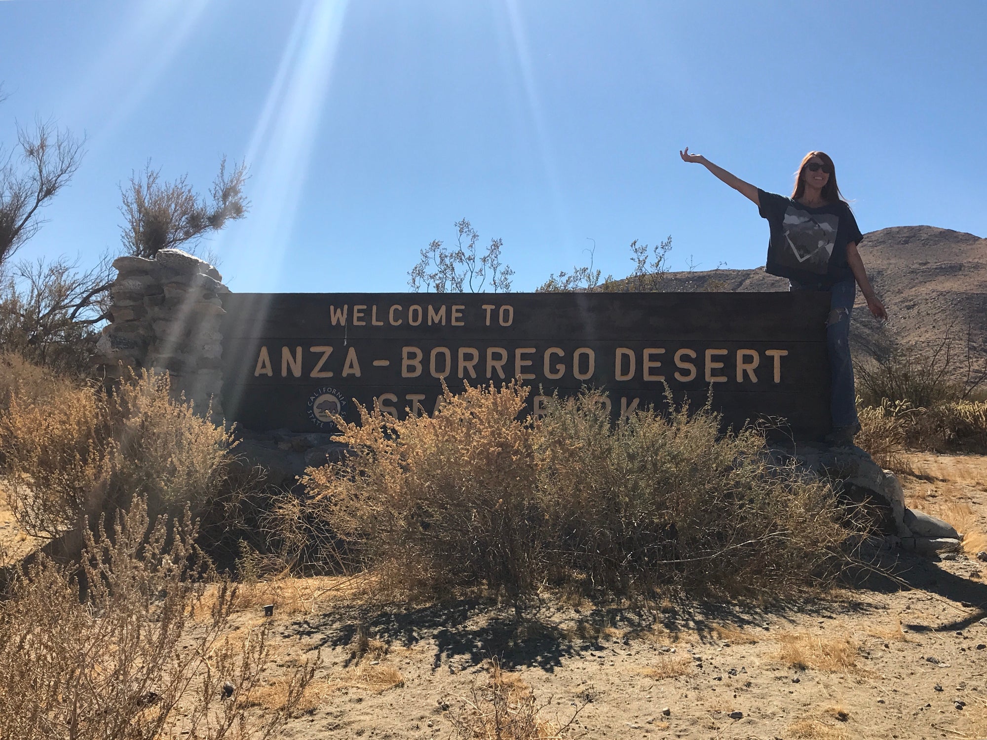 visitor poses beside the welcome sign to the woman poses for photo running away from a dinosaur sculpture in the anza-borrego desert
